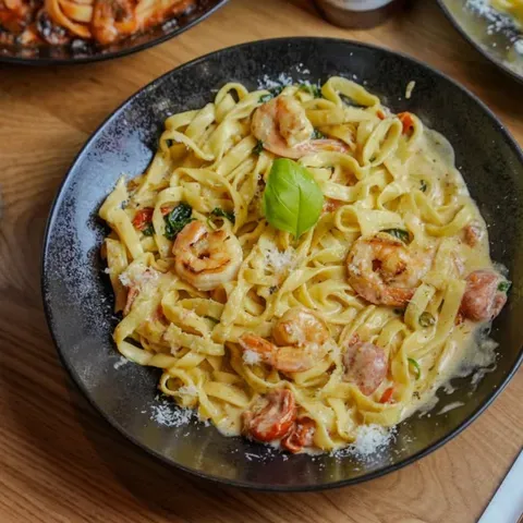 Eine Schüssel cremige Pasta mit Garnelen und Kräutern, garniert mit einem Basilikumblatt, auf einem Holztisch. Andere Gerichte sind teilweise um sie herum zu sehen.