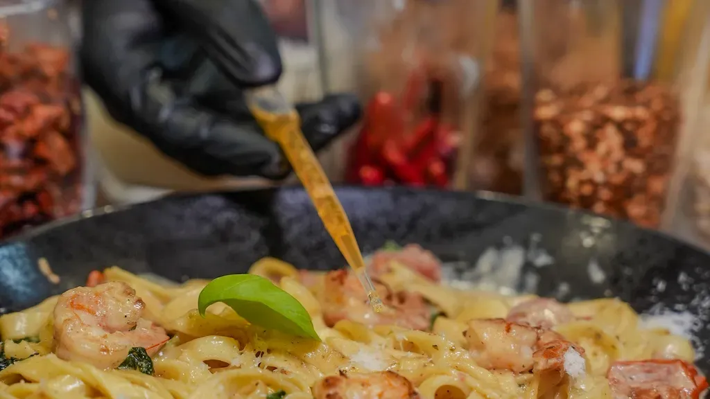Koch garniert cremige Garnelen-Pasta mit einer Pipette, umgeben von Gewürzen und Zutaten auf einem Holztisch.