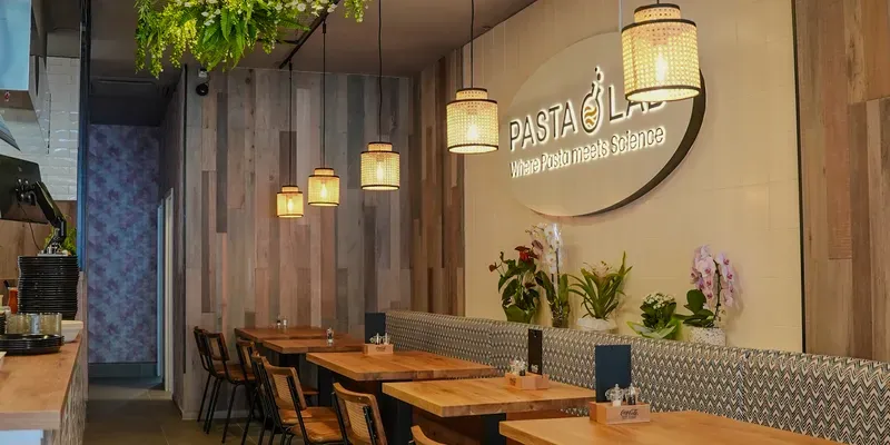 Gemütliches Restaurantinterieur mit Holztischen, schwarzen Stühlen, hängenden Pflanzen und warmer Beleuchtung. An der Wand hängt ein Schild mit der Aufschrift „PASTA“.