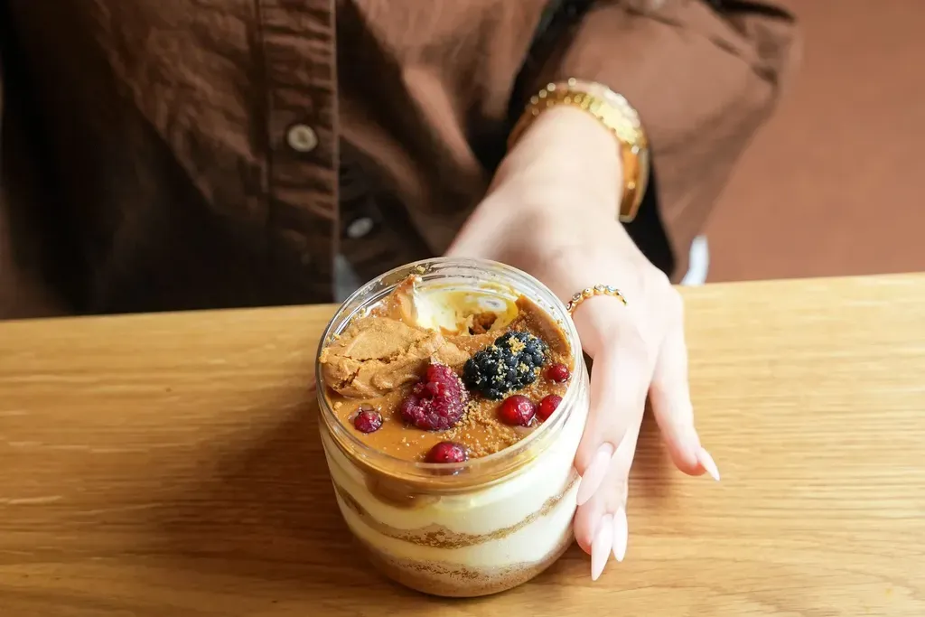 Eine Person in einem braunen Hemd, die ein Dessert in einem Glas mit Beeren darauf hält und an einem Holztisch sitzt.