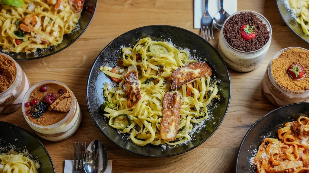 Ein Tisch mit verschiedenen Pastagerichten, Desserts mit Beeren und Schokolade sowie Besteck auf einer Holzplatte.