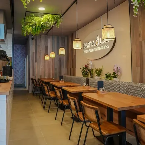 Gemütliches Restaurantinterieur mit Holztischen, Stühlen, hängenden Pflanzen und warmer Beleuchtung. An der gekachelten Wand hängt ein Schild mit der Aufschrift „PASTA“.