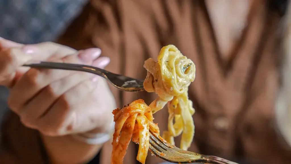 Frau hält Gabeln mit zwei Arten von Pasta, eine cremige und eine mit Tomatensoße, über einer Schüssel Pasta auf einem Holztisch.